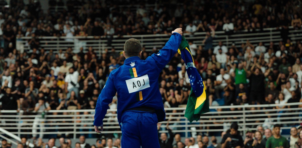 Tainan Dalpra, in an AOJ gi, raises the Brazilian flag to a packed arena after winning the 2026 Brasileiro